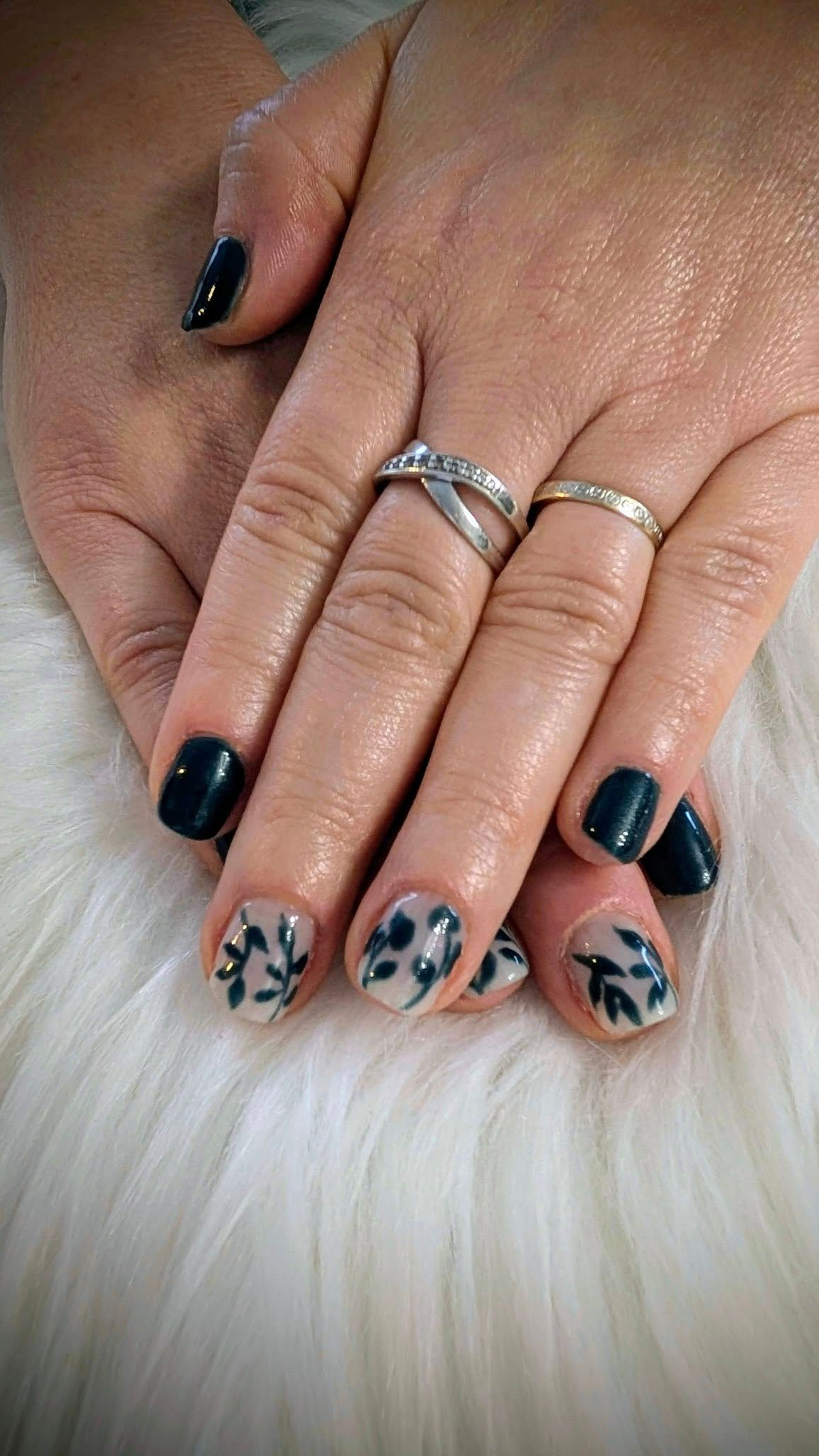 Des mains avec un vernis à ongles bleu foncé et un motif de feuille sur vernis transparent, posées sur une surface blanche et duveteuse.