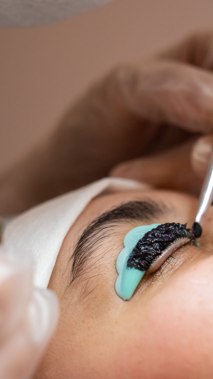 Une personne se fait faire un rehaussement de cils. Une main gantée applique une substance noire sur les cils à l'aide d'un pinceau, sur un moule bleu.