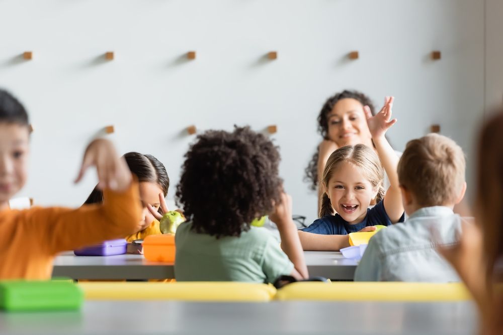 Niños y una maestra en un aula iluminada levantan la mano durante el almuerzo.