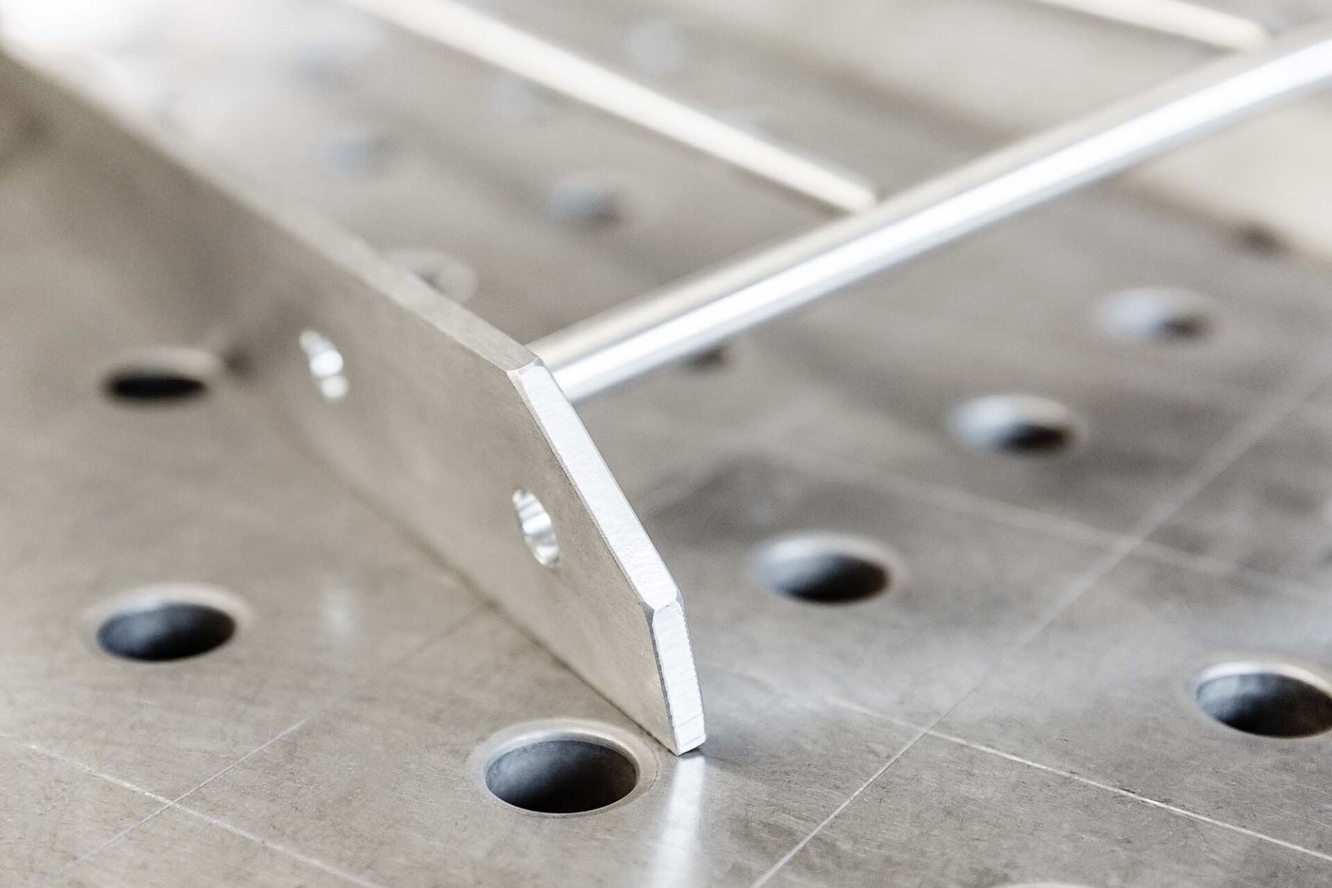 Nahaufnahme einer Metallkonstruktion, die eine leiterartige Struktur zeigt, die an ein flaches Stück geschweißt ist und auf einem Metalltisch mit Löchern ruht.
