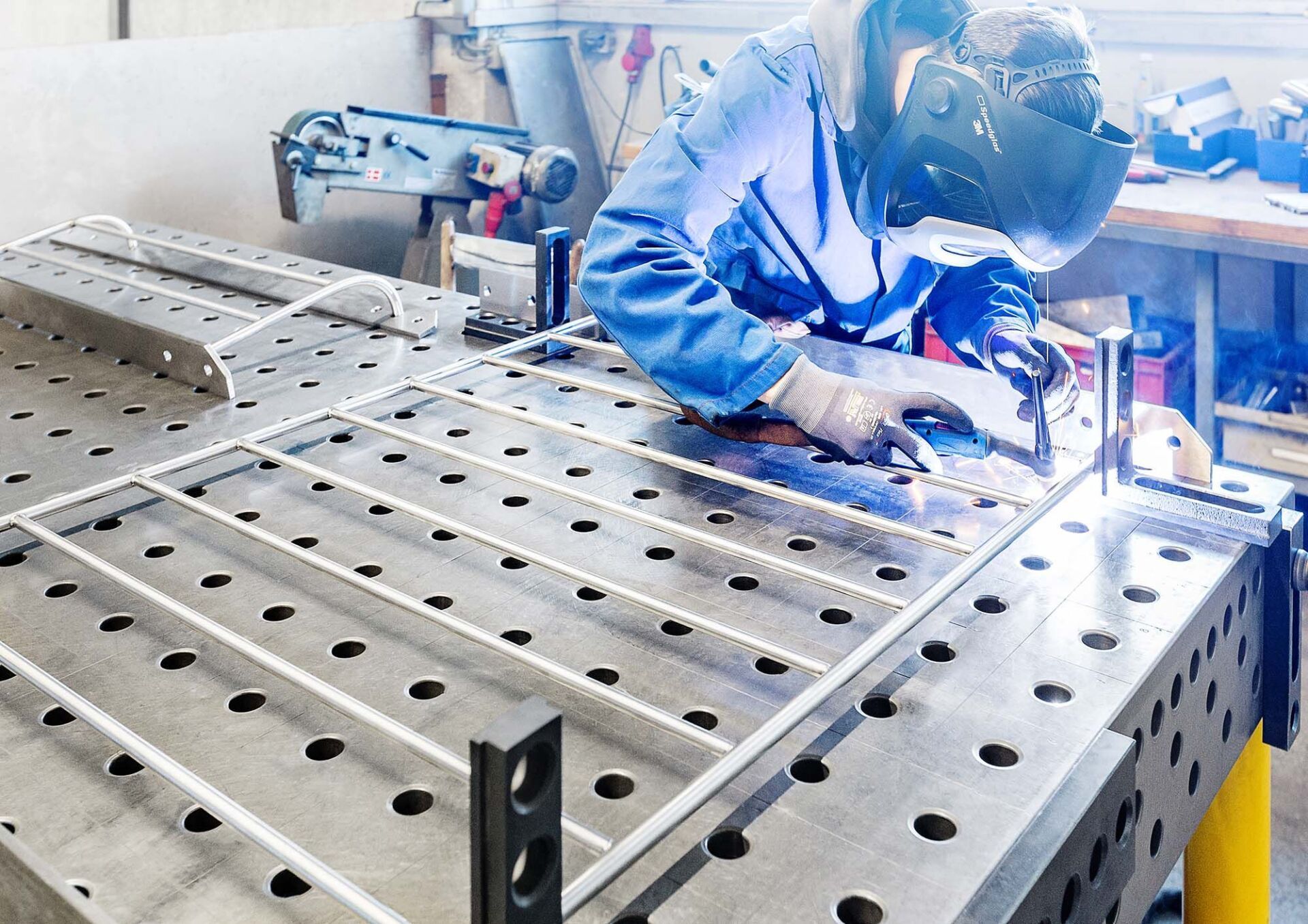 Schweißer in Schutzkleidung schweißt Metallrahmen auf einer perforierten Werkbank.