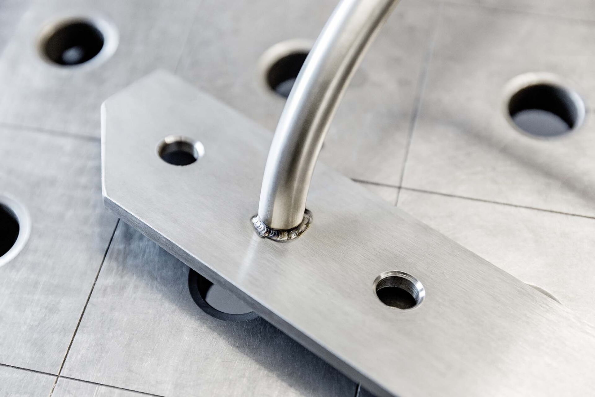 Nahaufnahme einer Metallhalterung mit drei Löchern, die an ein gebogenes Metallrohr geschweißt ist und auf einem Gittertisch steht.