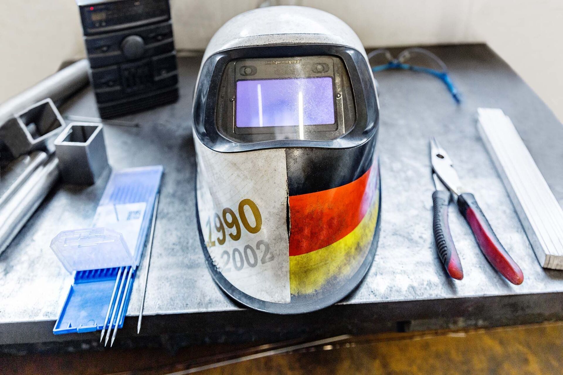 Schweißhelm mit deutscher Flagge, auf einem Metalltisch mit Werkzeugen und Zubehör.