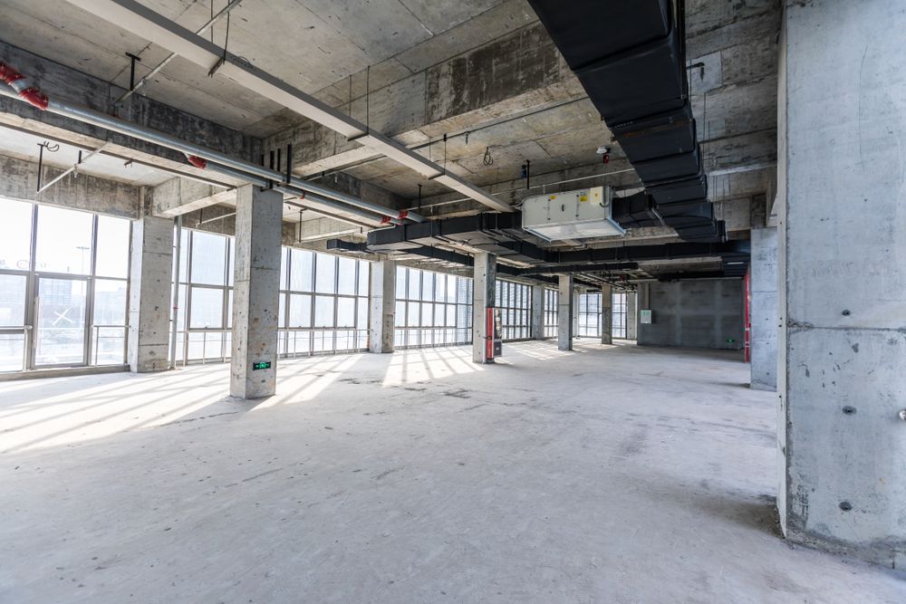 Interior de hormigón vacío sin terminar con grandes ventanas y servicios expuestos.