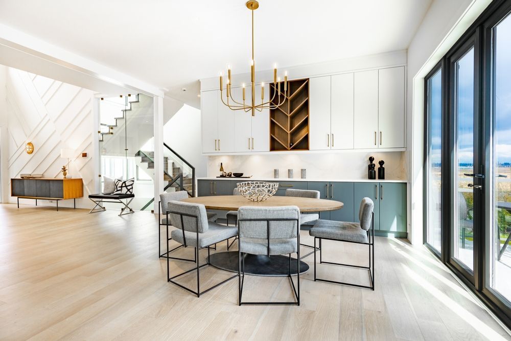 Comedor moderno con mesa ovalada, sillas grises, candelabro dorado, grandes ventanales.