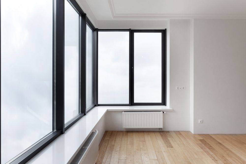 Habitación vacía con grandes ventanales, suelo de madera clara, paredes blancas y radiador.
