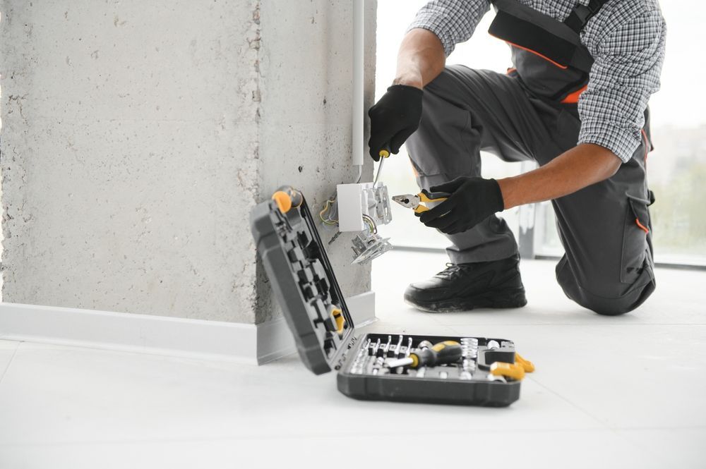 Electricista instalando un enchufe. Lleva guantes negros, ropa de trabajo gris y botas negras. Caja de herramientas abierta en el suelo.