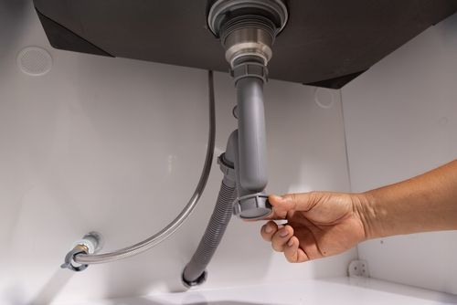 Una persona desmonta el sifón de un fregadero blanco. Se ven tuberías grises y una manguera flexible.