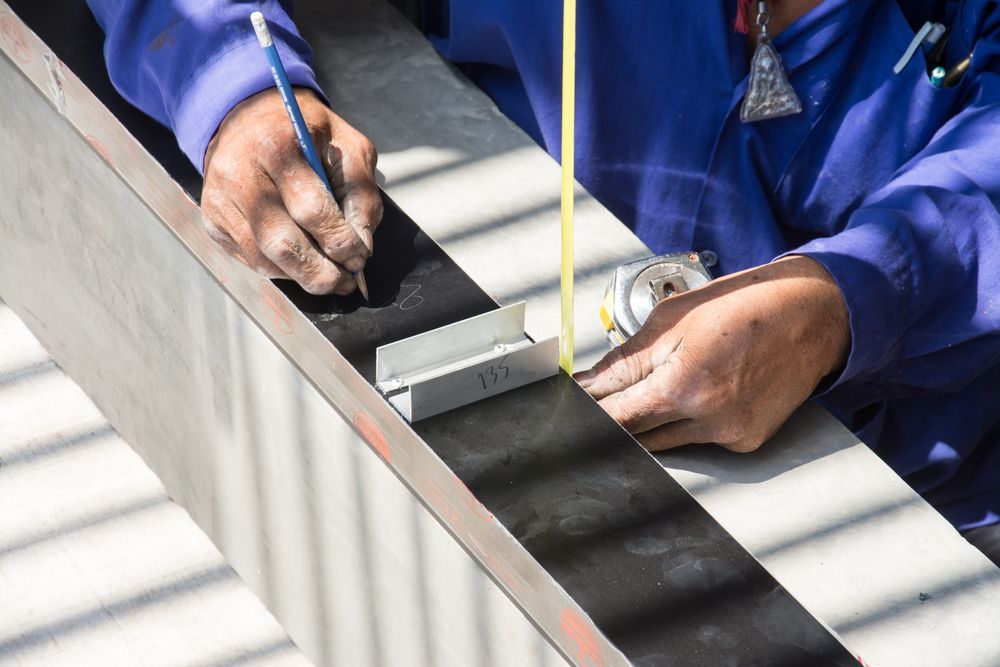 Persona con mono azul que utiliza un lápiz y una cinta métrica para marcar una viga de metal.