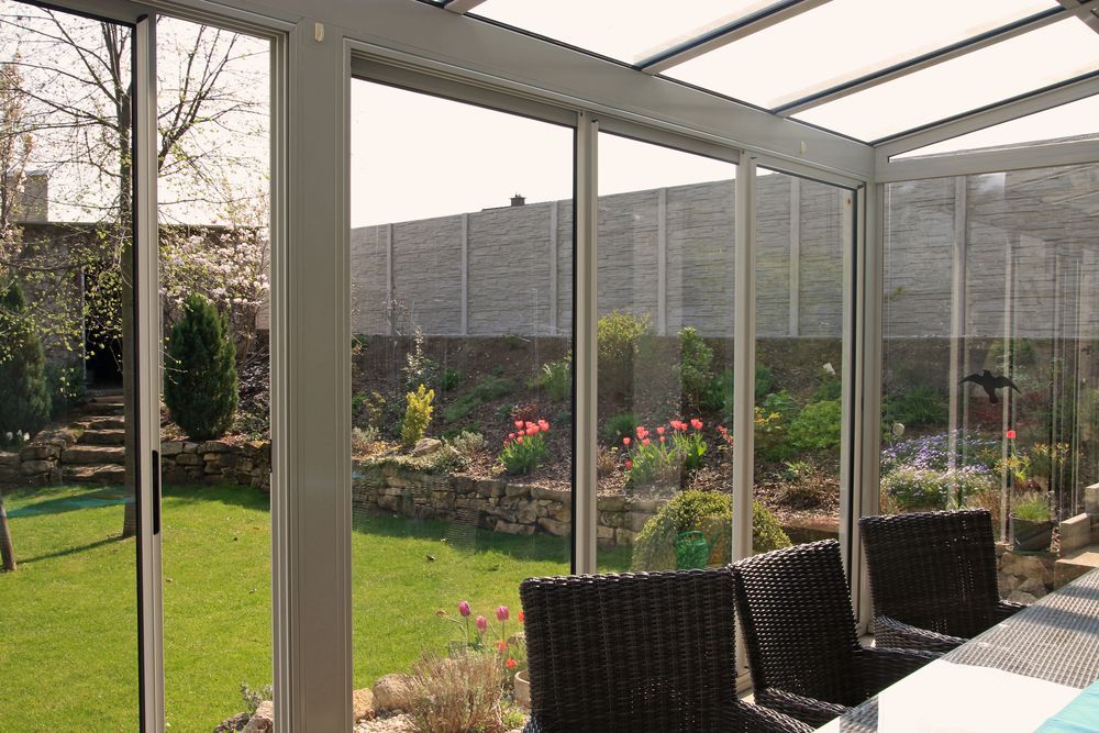 Interior de terraza acristalada con vista a un jardín; sillas de mimbre en una mesa, altos ventanales de vidrio, césped verde.