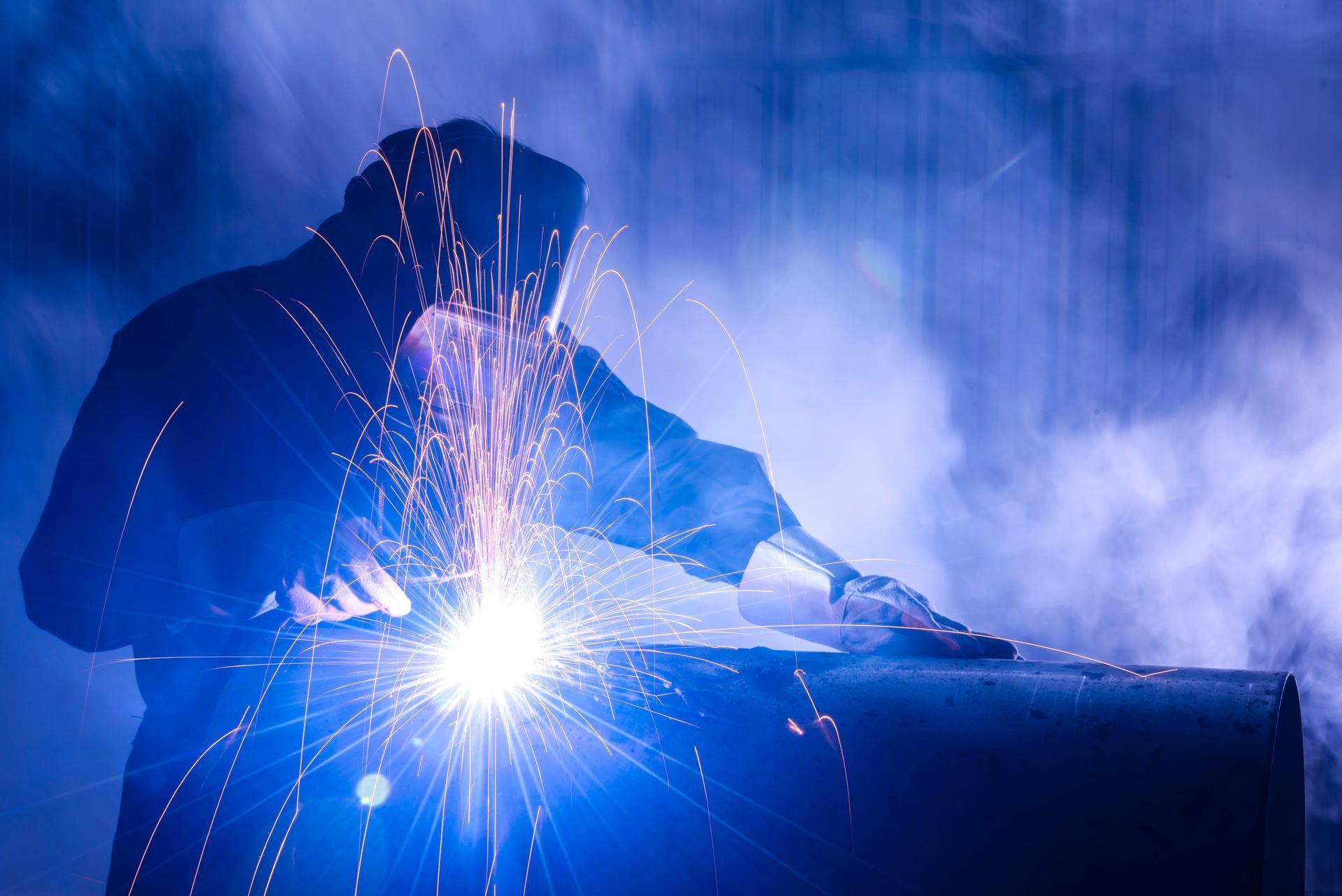 Soudeur en vêtements sombres travaillant sur du métal, étincelles jaillissant, ambiance bleutée.