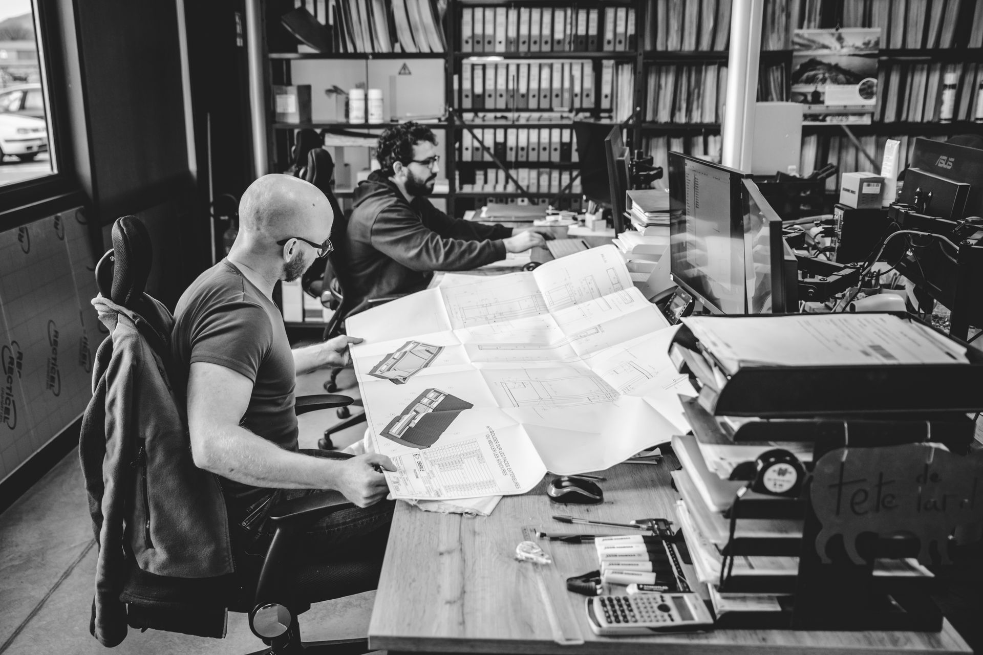 Deux hommes dans un bureau examinent une feuille de papier pliée ; l'un d'eux est assis à un bureau avec un ordinateur