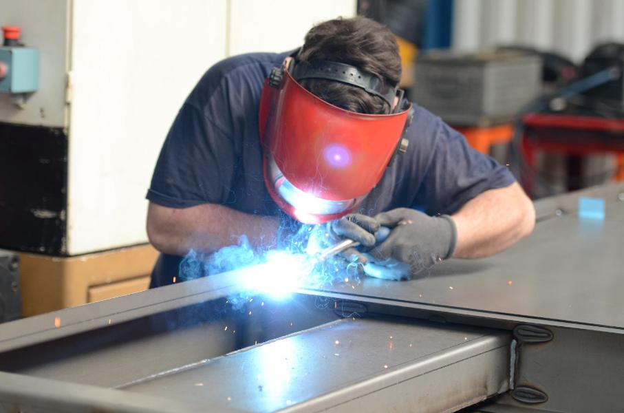 Un soudeur portant un masque rouge travaille sur du métal, projetant des étincelles  dans un environnement industriel
