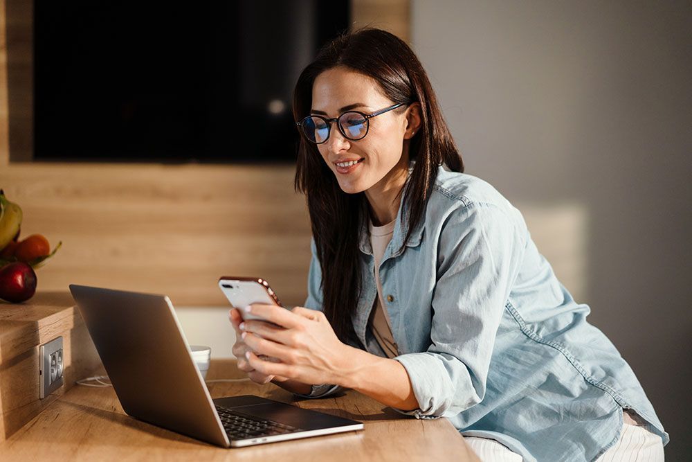Eine Frau sitzt an einem Tisch und benutzt einen Laptop und ein Mobiltelefon.