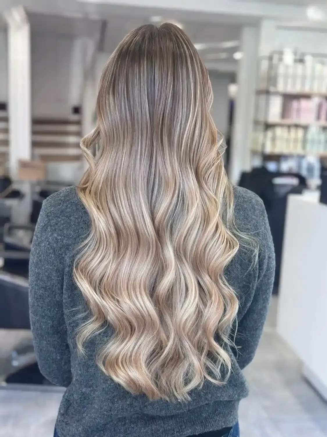 Une personne vue de dos, montrant de longs cheveux blonds ondulés avec des mèches, portant un pull gris dans un salon de coiffure.