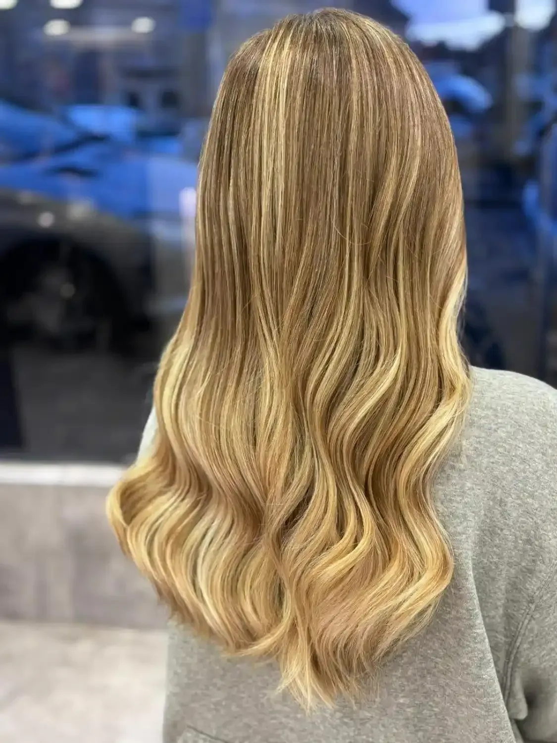 Une personne vue de dos, aux longs cheveux blonds ondulés avec des mèches plus claires, coiffés en ondulations souples.