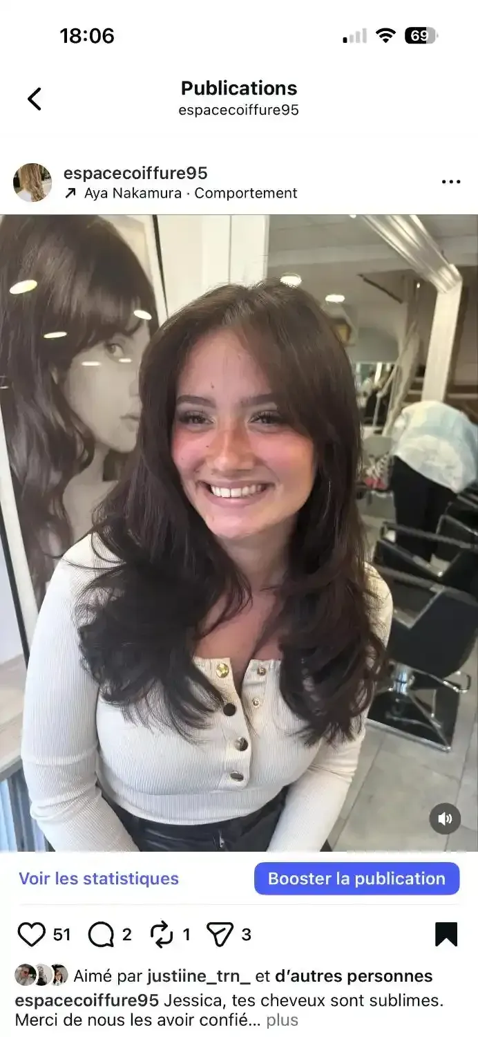 Une personne souriante aux cheveux foncés, dégradés et mi-longs, portant un haut blanc, dans un salon de coiffure.