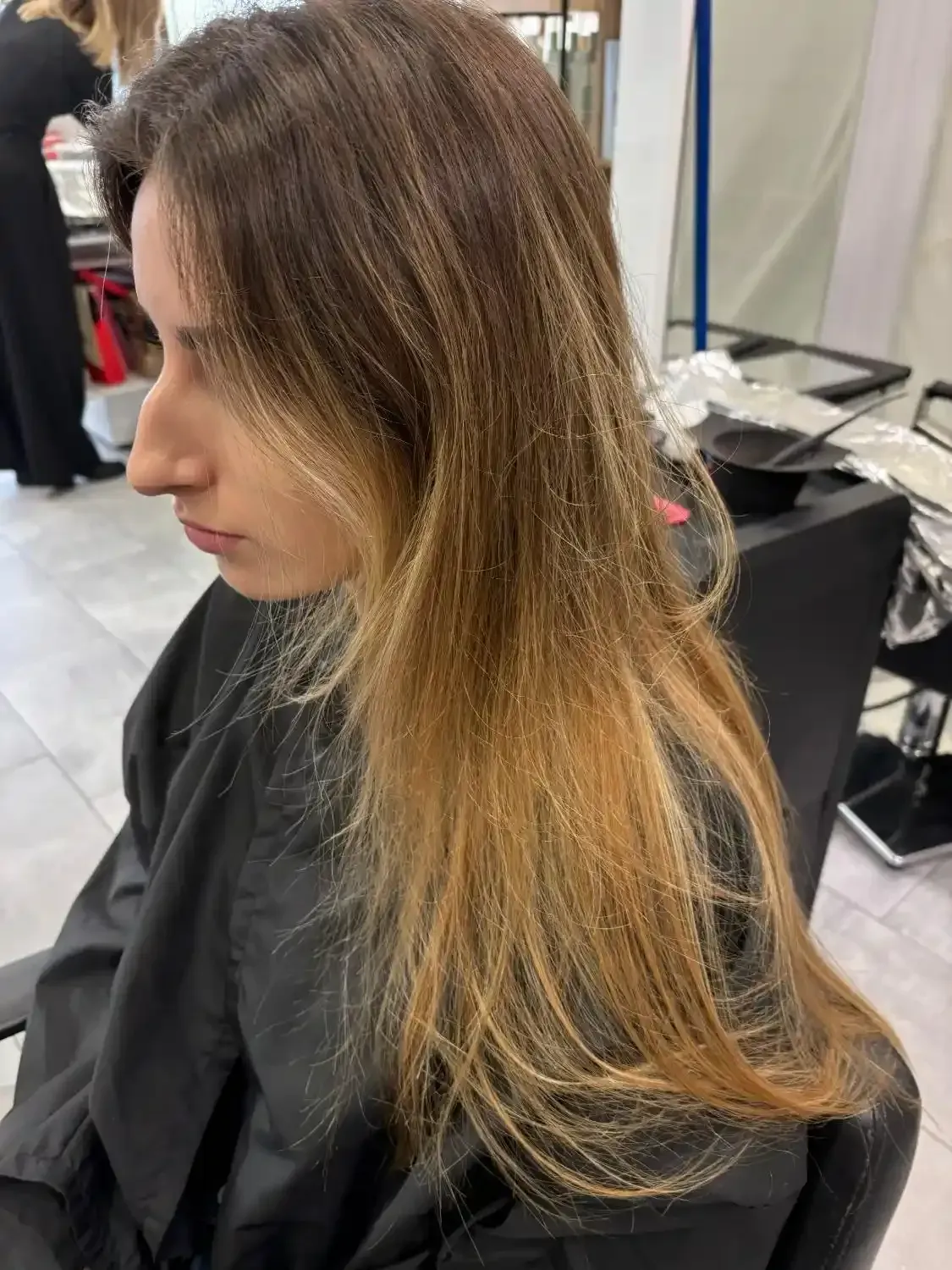 Vue de profil d'une personne assise dans un fauteuil de salon, aux longs cheveux bruns méchés qui s'éclaircissent jusqu'à un blond clair aux pointes.