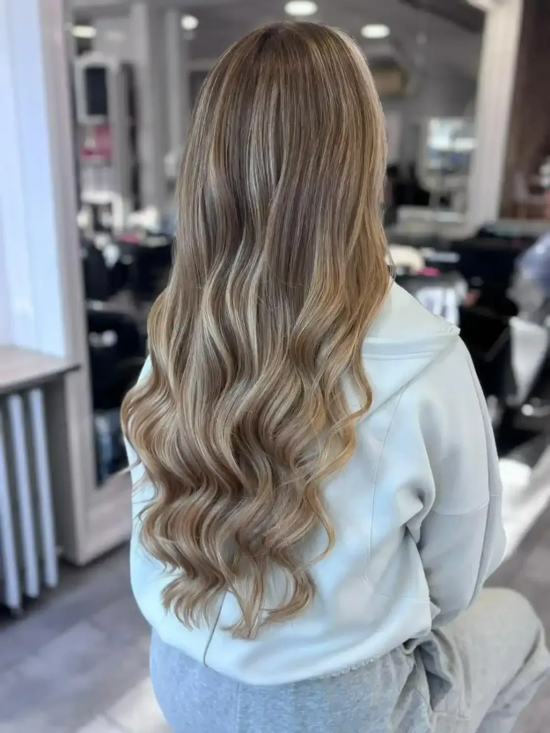 Une personne aux cheveux longs, ondulés et châtain clair méchés, vue de dos, portant un pull gris clair dans un salon de coiffure.
