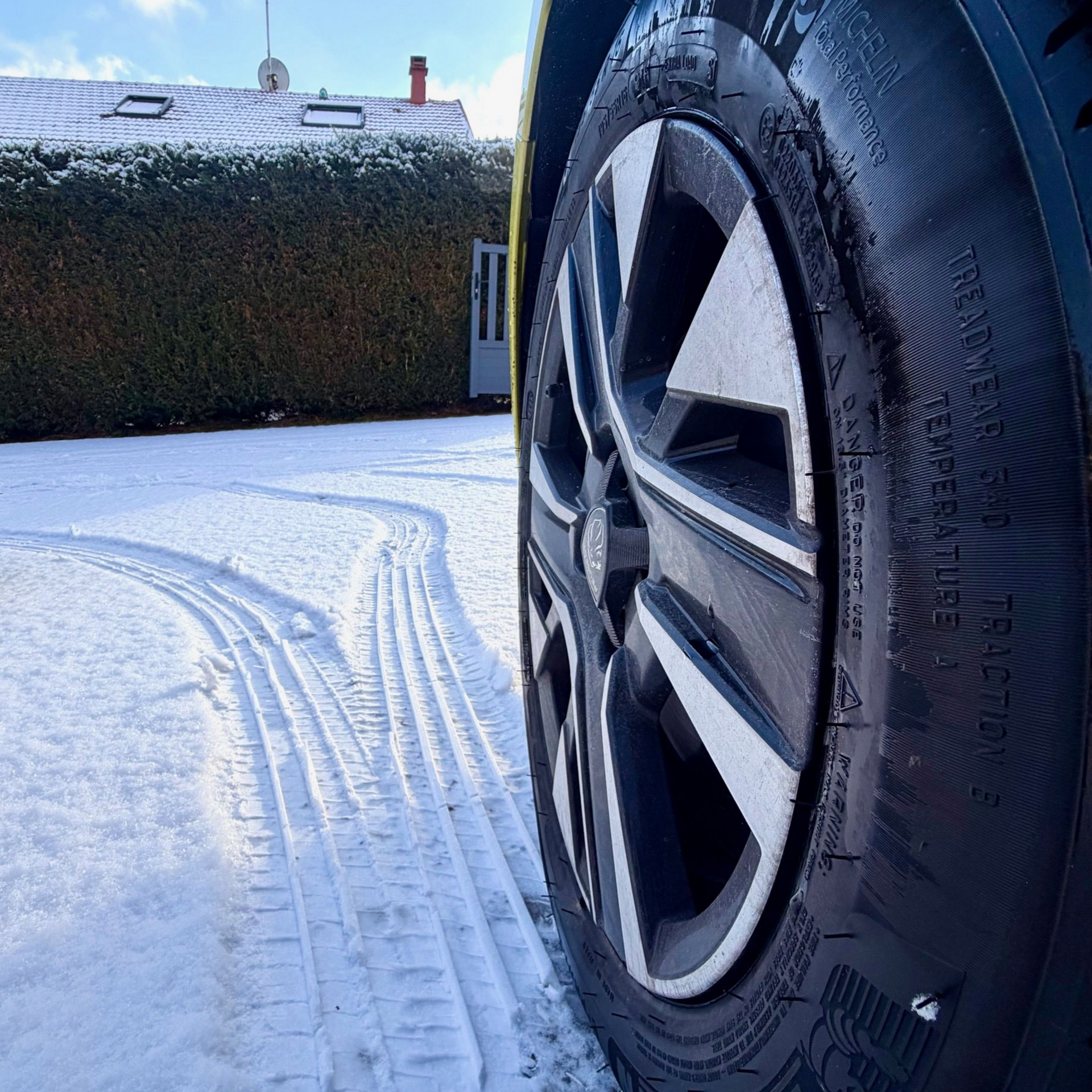 Pneu de voiture dans la neige, laissant des traces. Le pneu est noir avec une jante argentée. La neige recouvre le sol et une haie verte se dresse en arrière-plan.