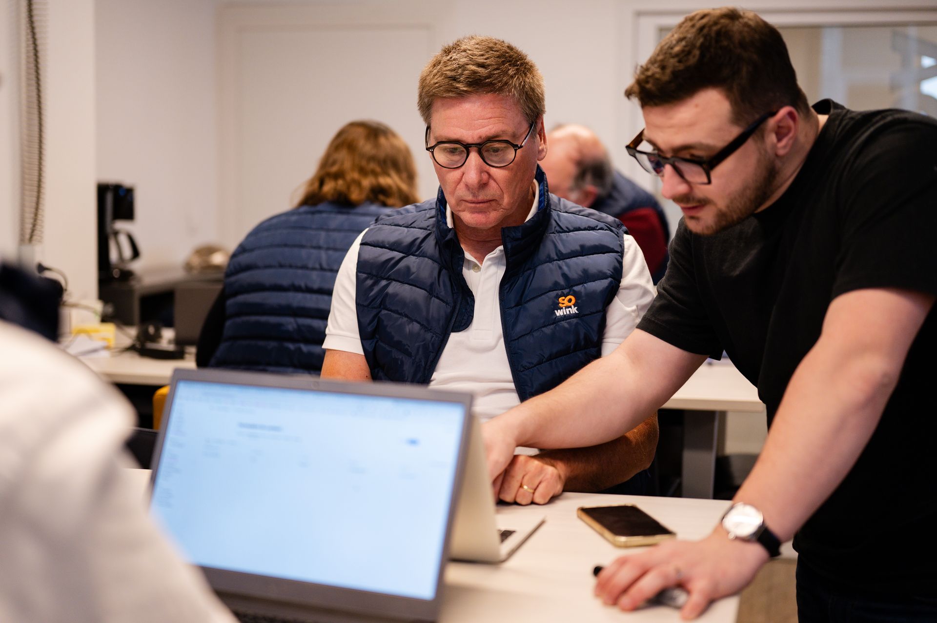 Deux personnes collaborent sur un ordinateur portable dans un bureau, l'une d'elles faisant des gestes vers l'écran.