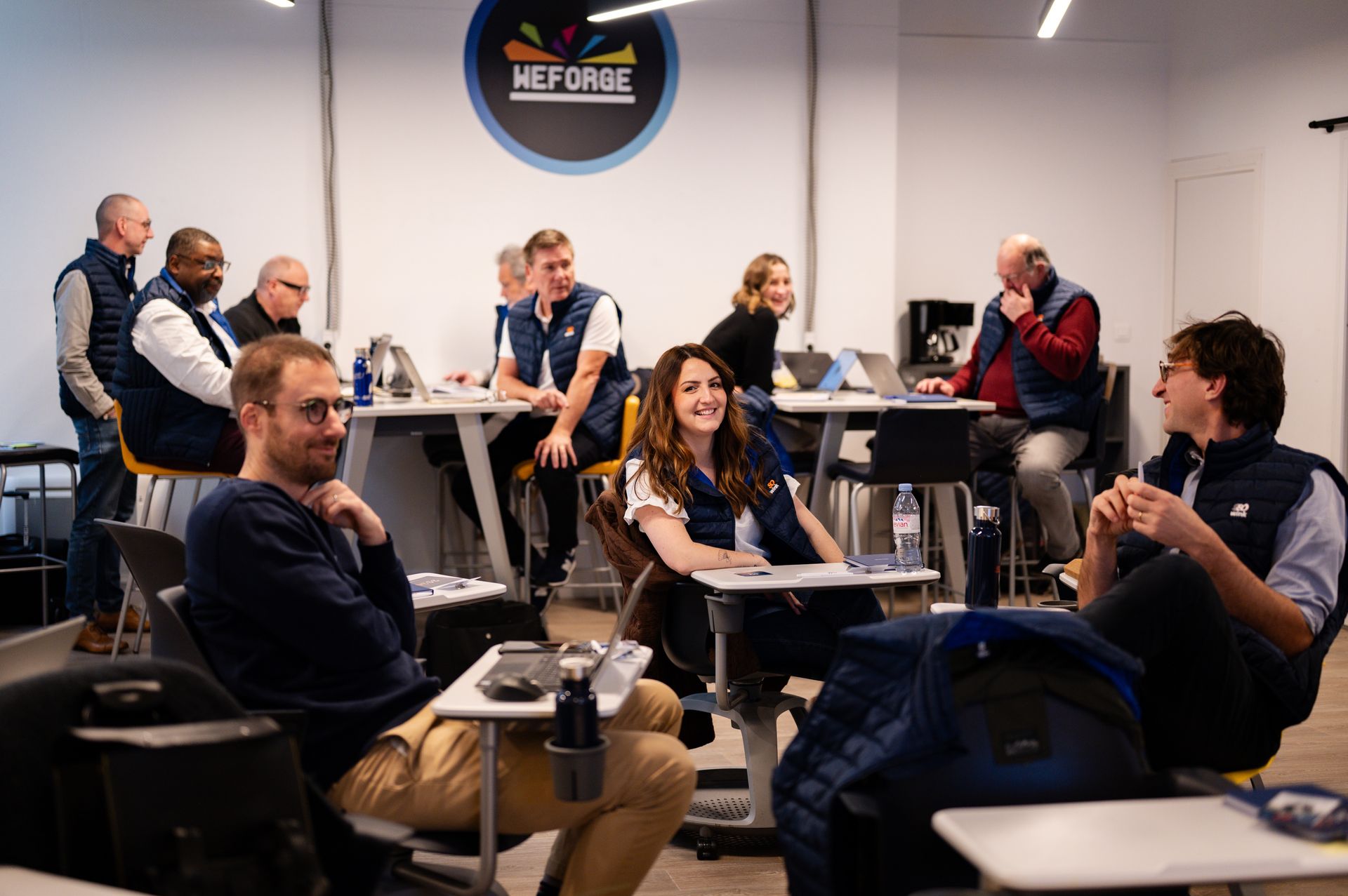 Dans un espace de travail, un groupe de personnes est assis à des bureaux et des tables ; certaines discutent, d'autres travaillent sur des ordinateurs portables.