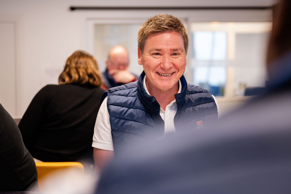 Dans une pièce lumineuse, une personne vêtue d'une chemise blanche et d'un gilet bleu marine sourit tandis que d'autres personnes sont assises à l'arrière-plan.