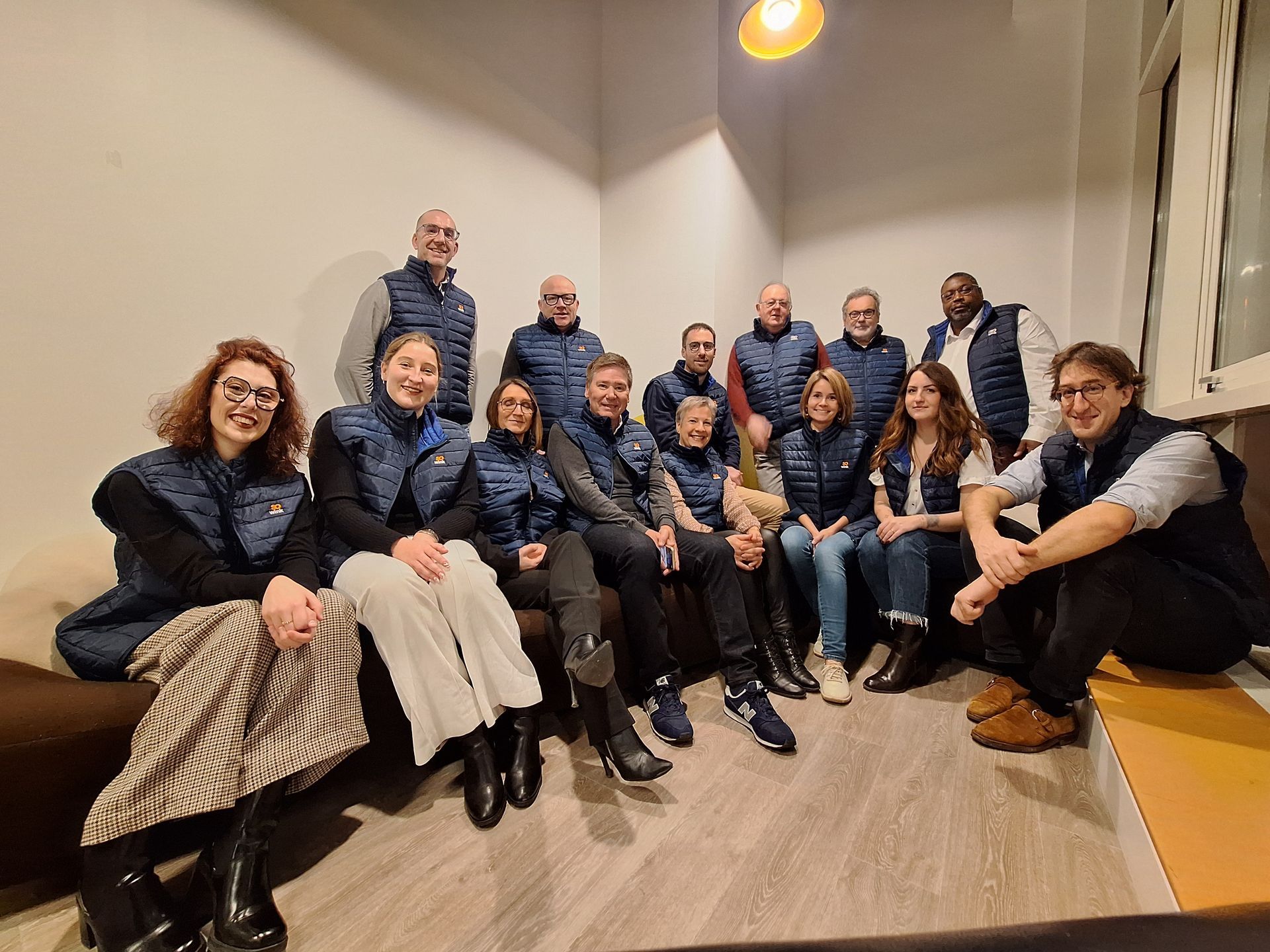 Un groupe de quinze personnes vêtues de gilets bleus assortis pose ensemble dans un espace de bureaux intérieur lumineux.