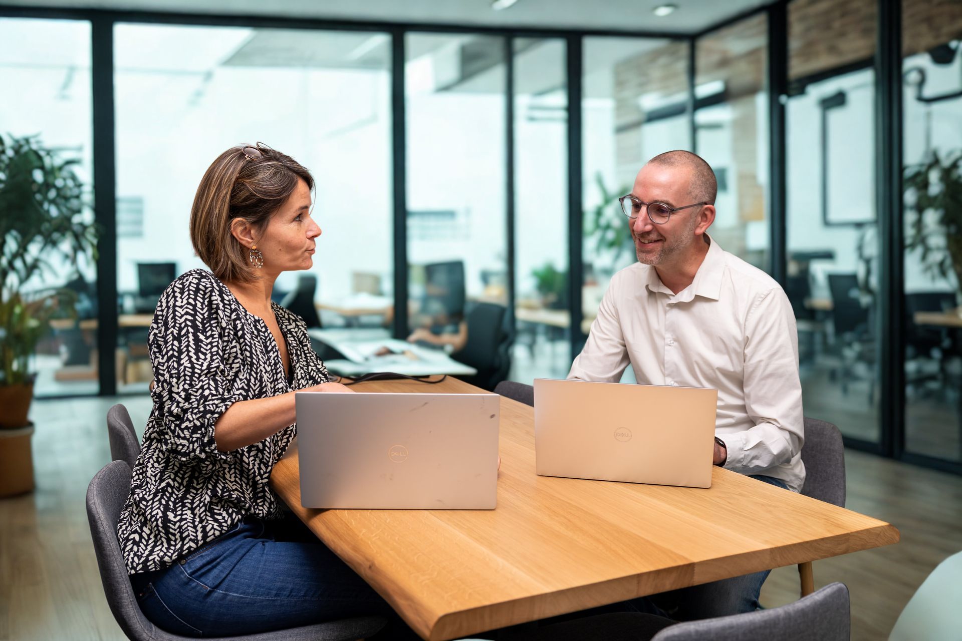Dans un bureau, une femme et un homme sont assis l'un en face de l'autre à une table en bois, chacun travaillant sur son propre ordinateur portable.