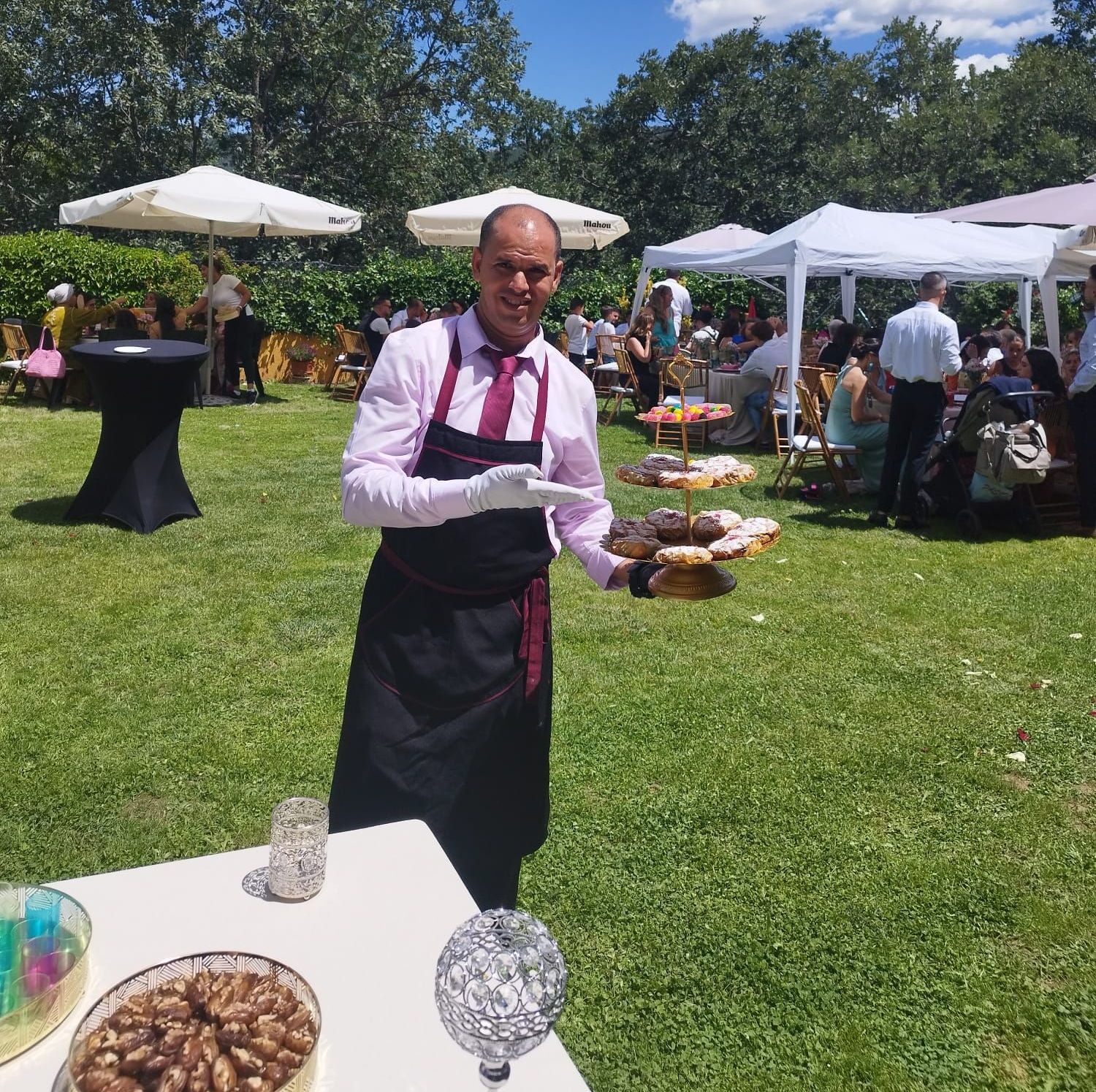 Camarero sosteniendo una bandeja de comida al aire libre en un evento soleado, otros invitados sentados en mesas en el fondo.