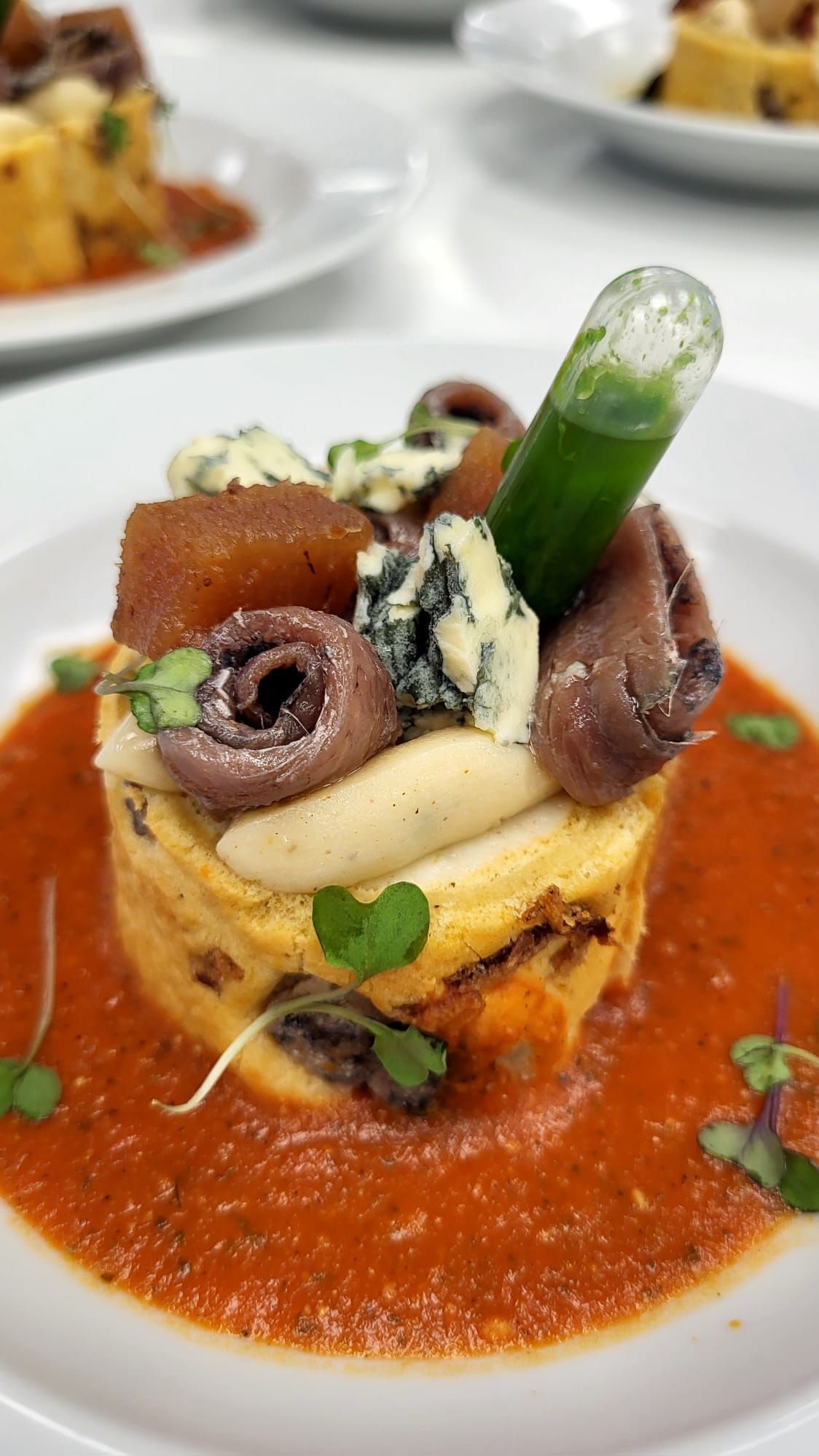 Plato gourmet: Polenta apilada, anchoas, queso azul, salsa de tomate y un elemento verde.