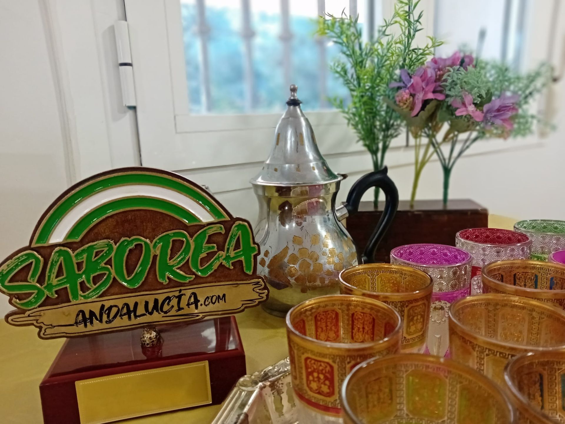 Logotipo de SABOREA ANDALUCIA junto a vasos de té y una tetera plateada, cerca de una ventana con flores.