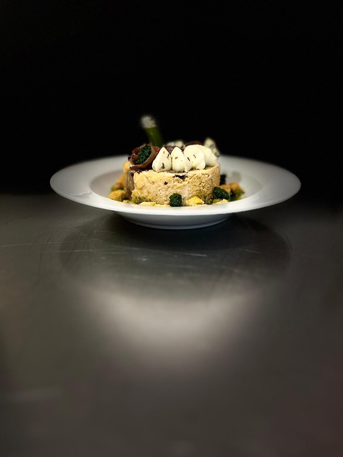 Plato blanco con un plato gourmet. El plato reposa sobre una superficie reflectante. La comida tiene componentes de color crema claro y guarniciones verdes.