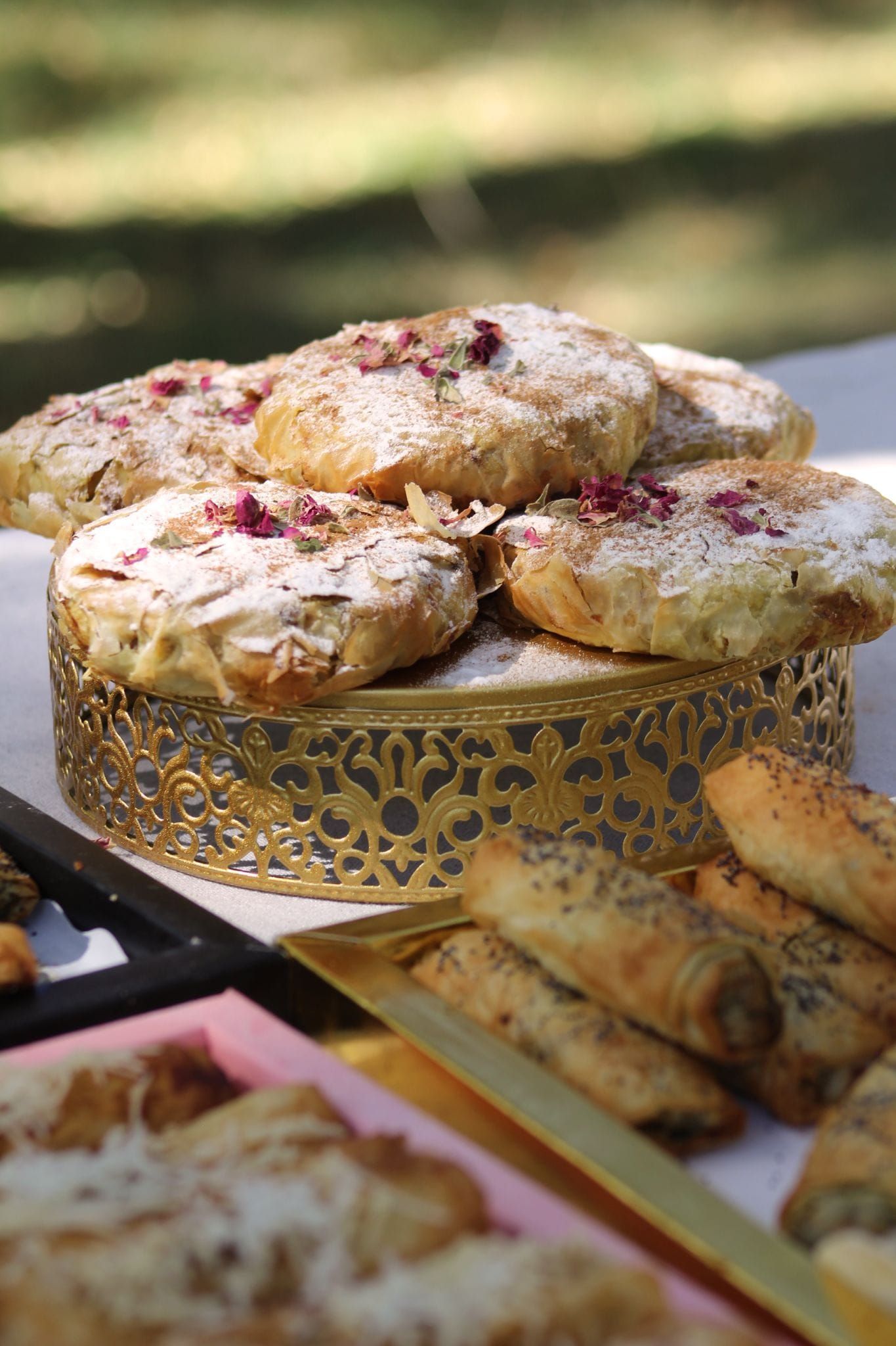 Los pasteles, incluidos los redondos espolvoreados con azúcar glas y los panecillos, se disponen en una bandeja de servicio dorada al aire libre.