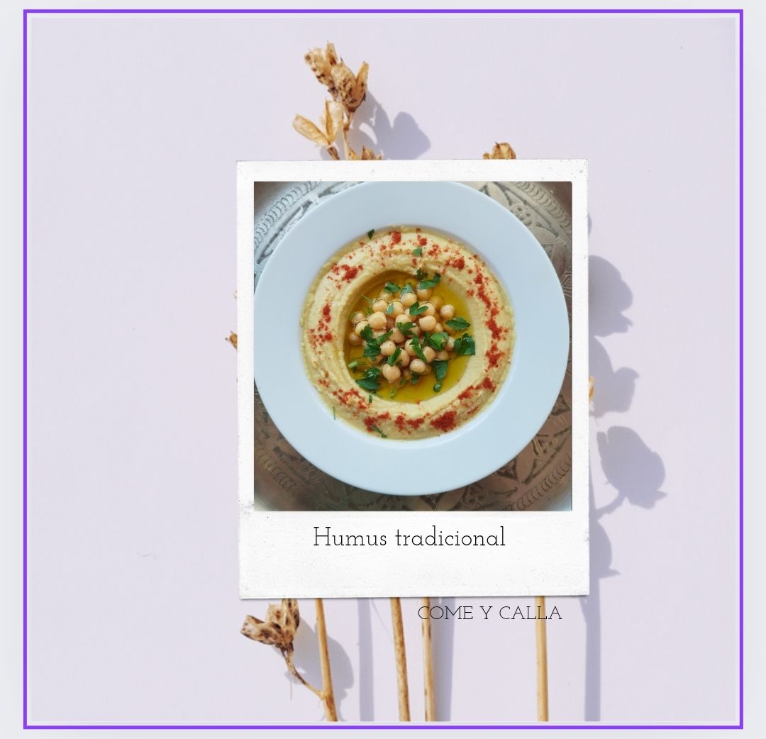 Una imagen de un tazón de hummus con garbanzos sobre un fondo morado.