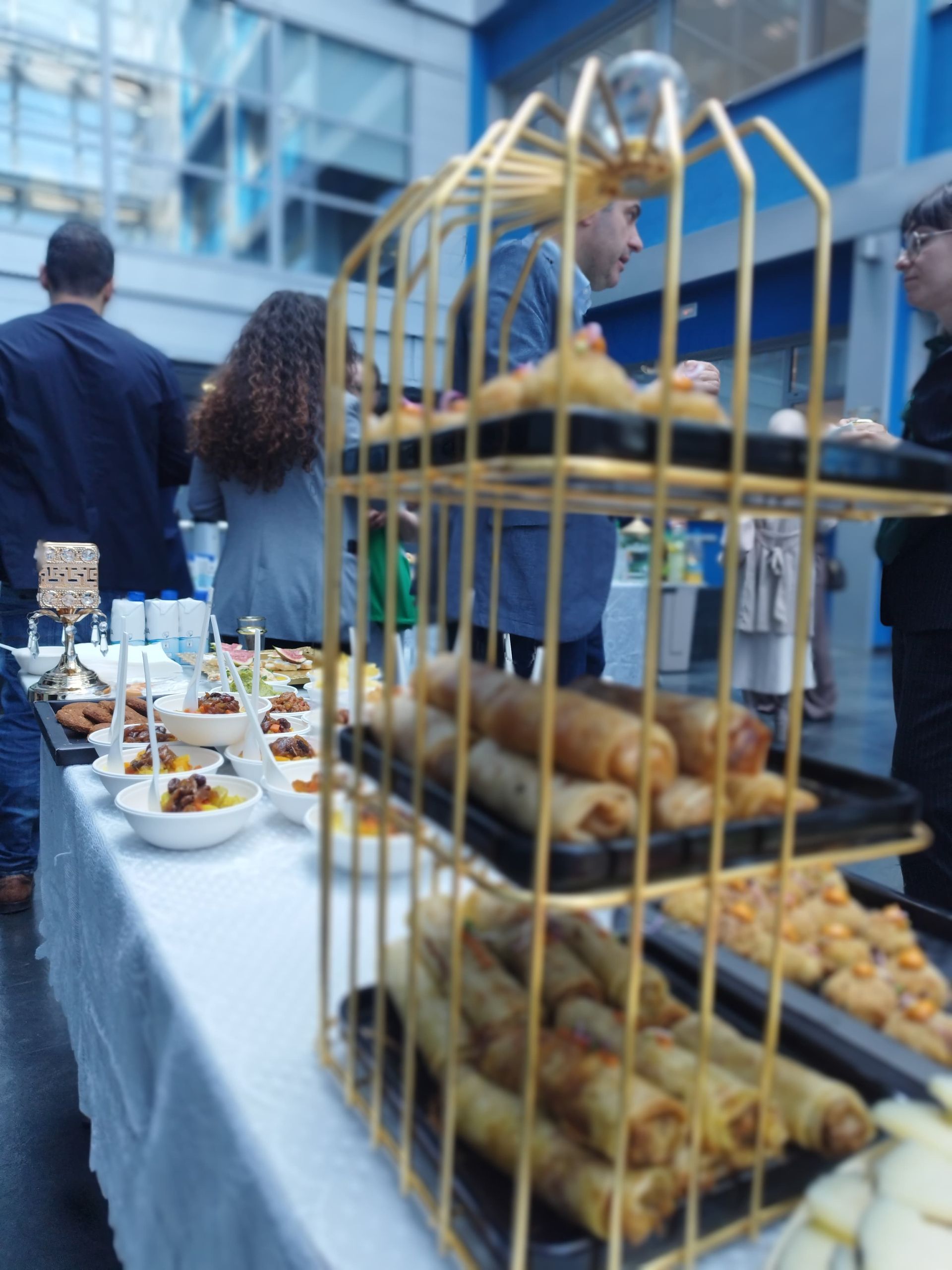 Mesa de buffet con comida en bandejas, rejilla de metal dorado, gente de fondo.
