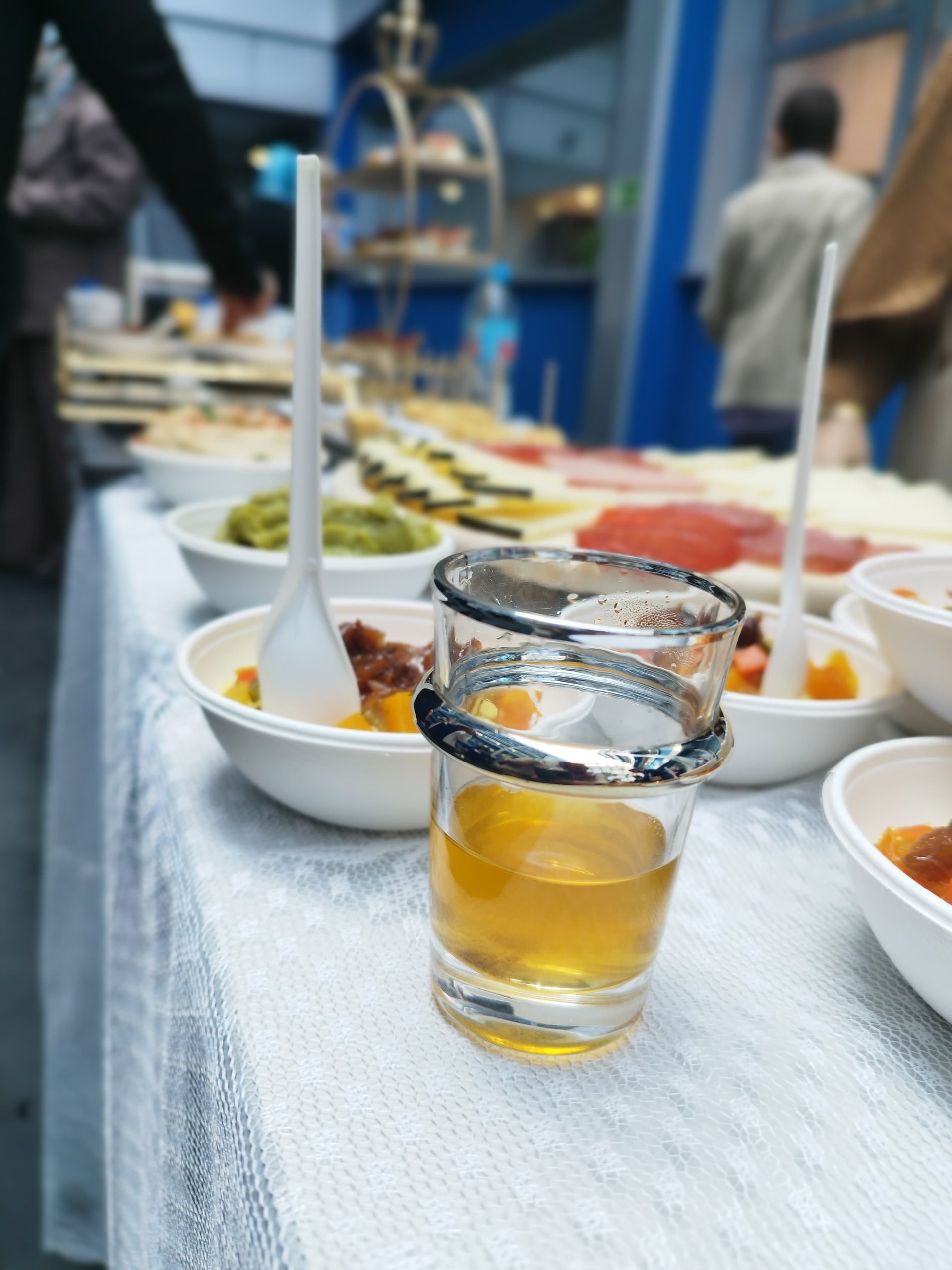 Un vaso de chupito con líquido ámbar sobre una mesa llena de comida. Otras personas al fondo.