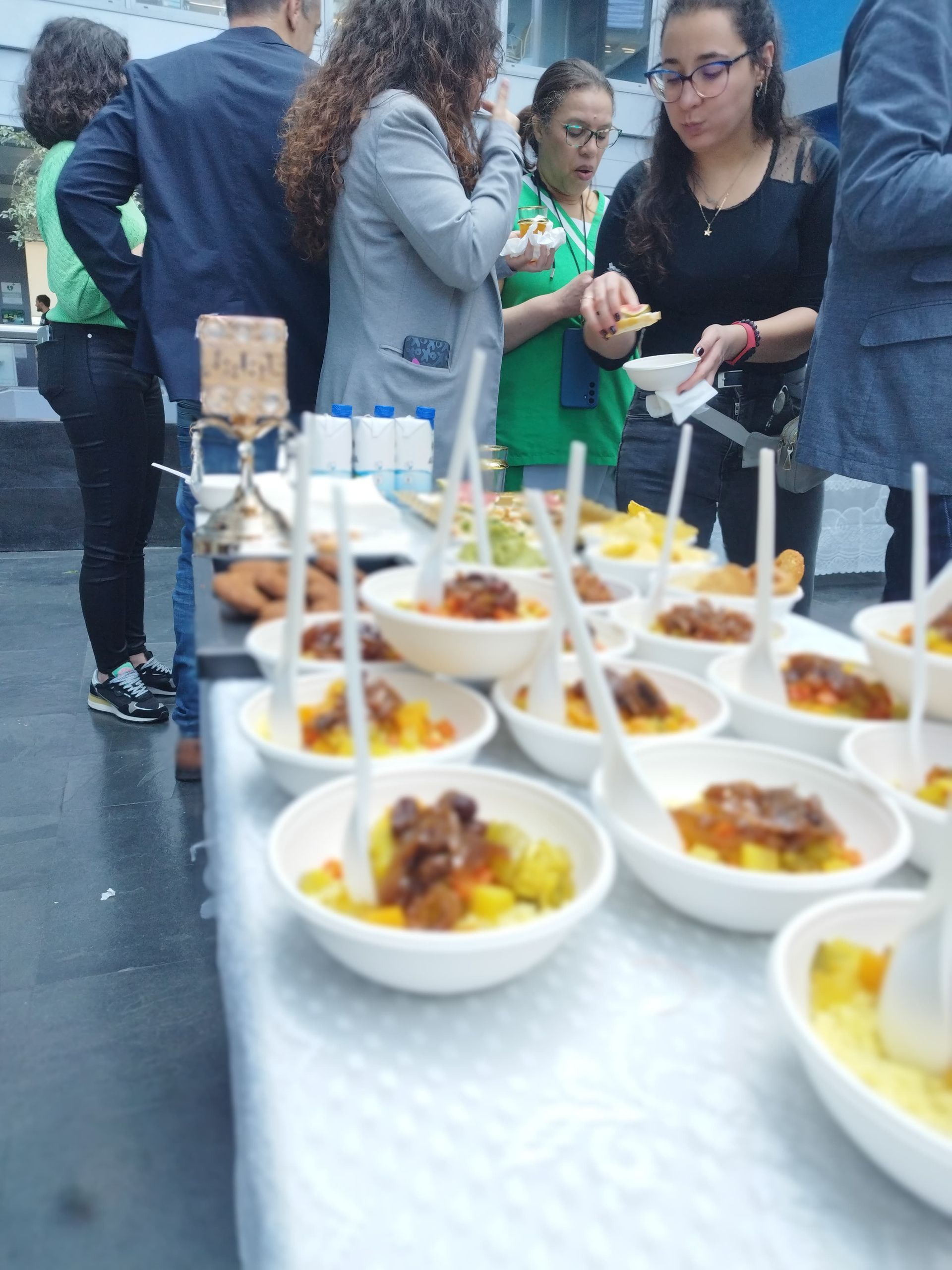 Personas comiendo en pequeños tazones en un evento al aire libre. Comida en una mesa con cucharas para servir.