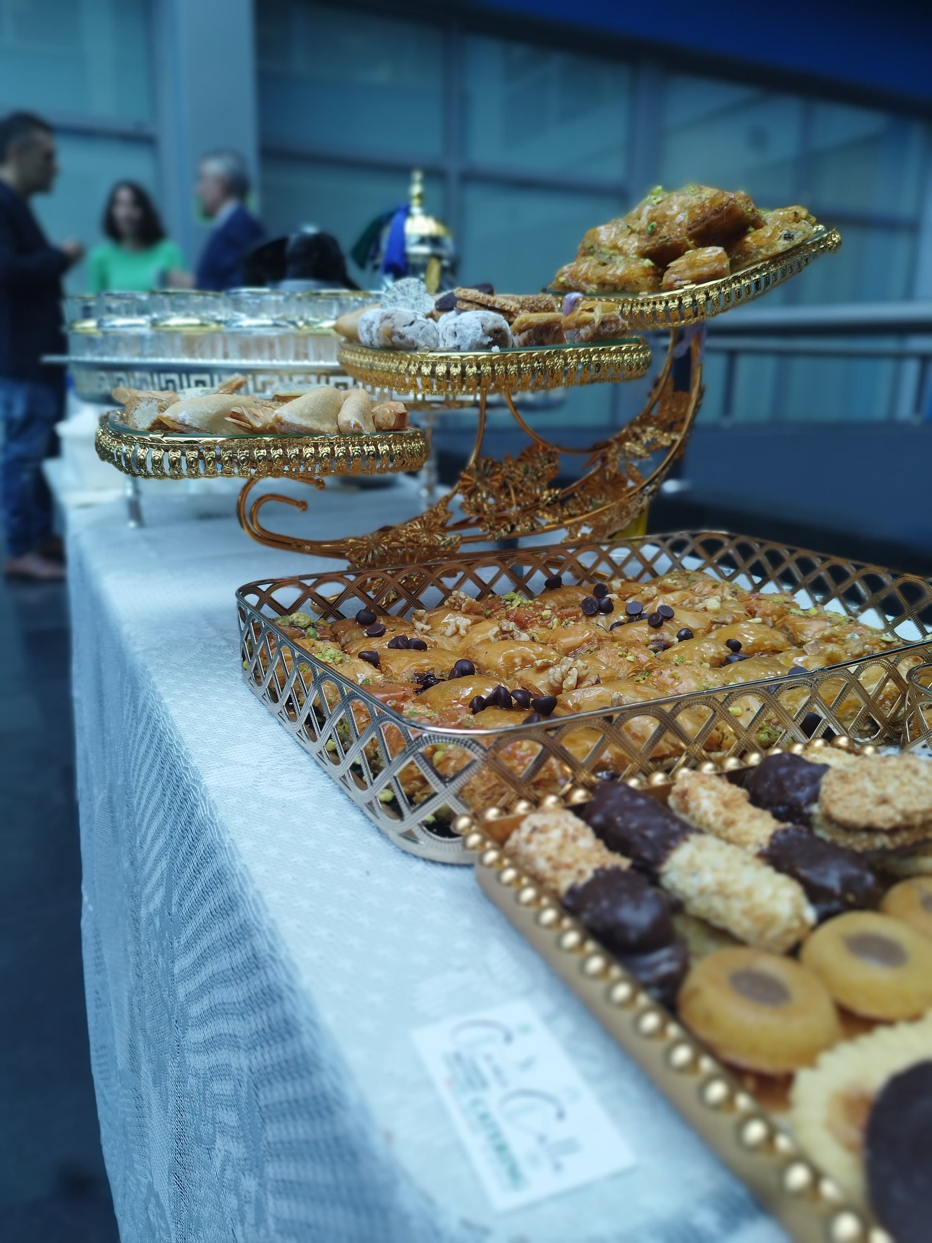 Galletas y pasteles en bandejas escalonadas durante un evento. Gente de pie al fondo.