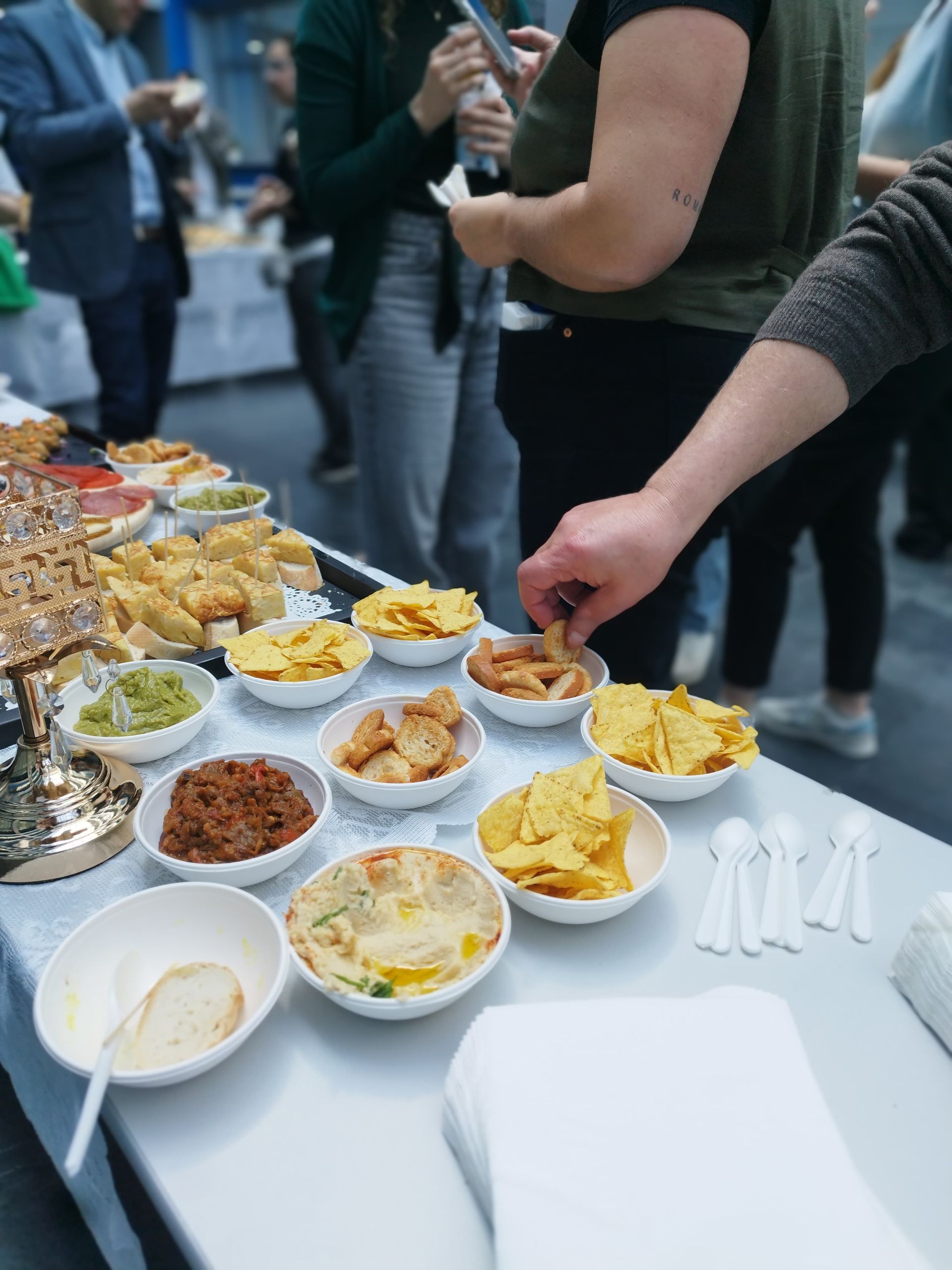 Mesa de buffet con diversas salsas y bocadillos, gente buscando comida.