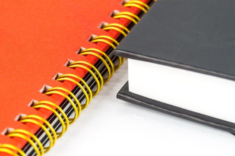 Orangenes Ringbuch mit gelben Ringen, das neben einem blau eingebundenen Buch liegt.