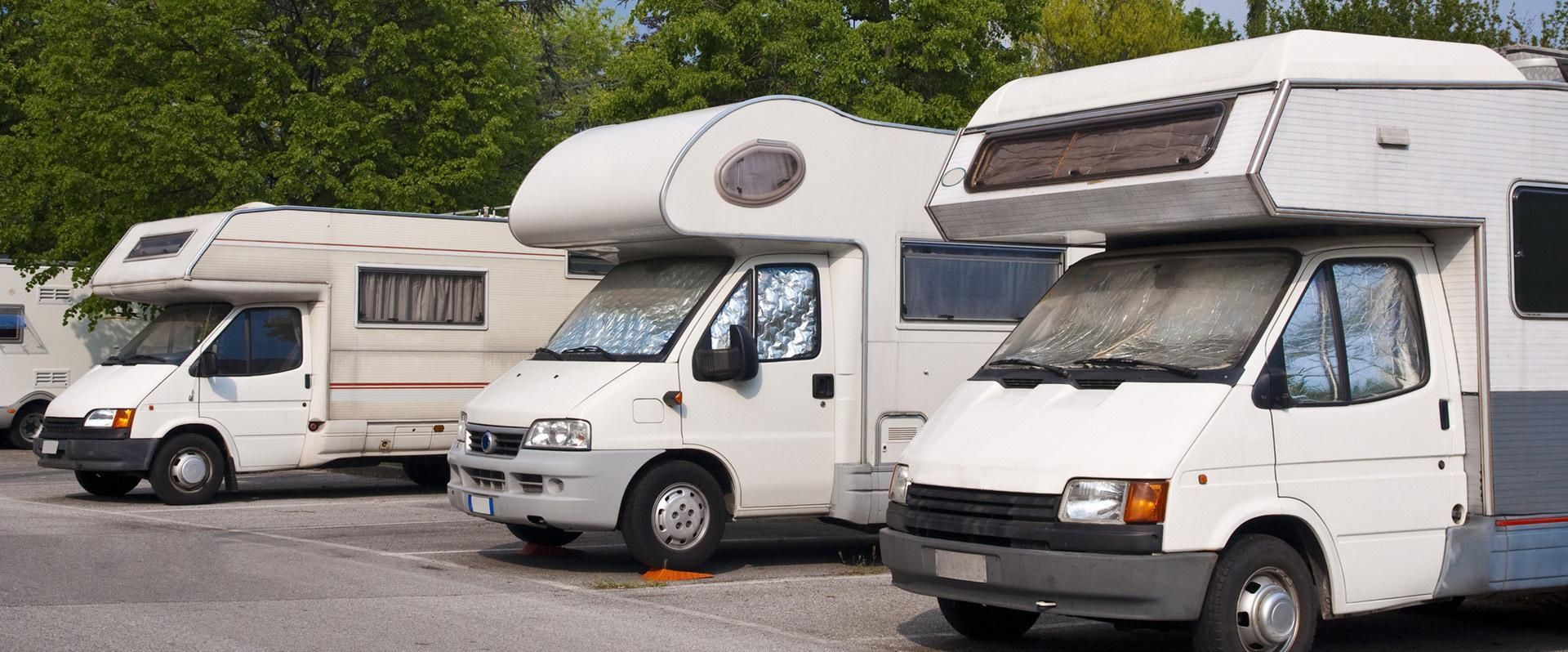 Des camping-cars blancs stationnés sur un parking en gravier, à côté d’arbres