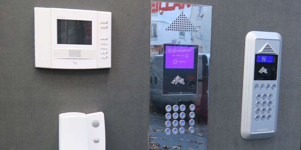 Gros plan sur un interphone et son panneau de commande fixés sur un mur gris. On y voit un écran, un clavier et des boutons.