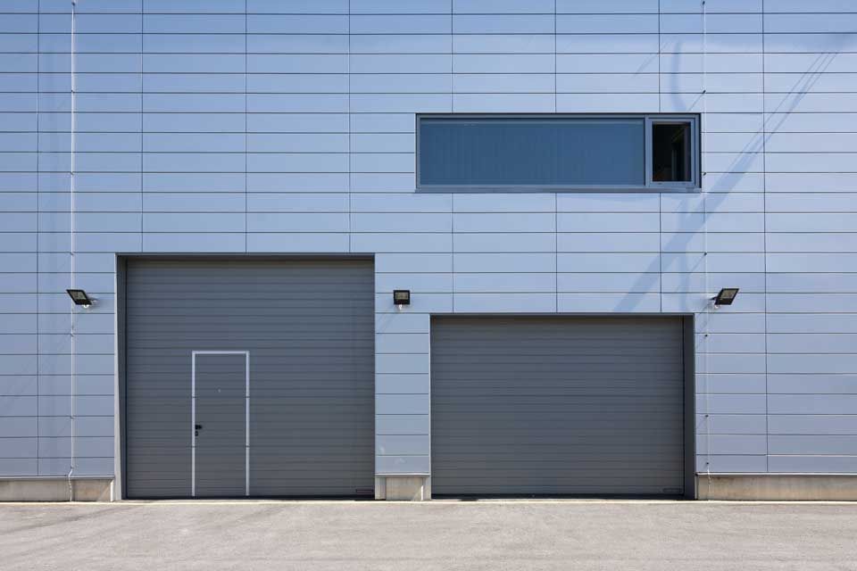 L'extérieur d'un bâtiment moderne avec deux portes de garage grises, une petite porte et une fenêtre rectangulaire.
