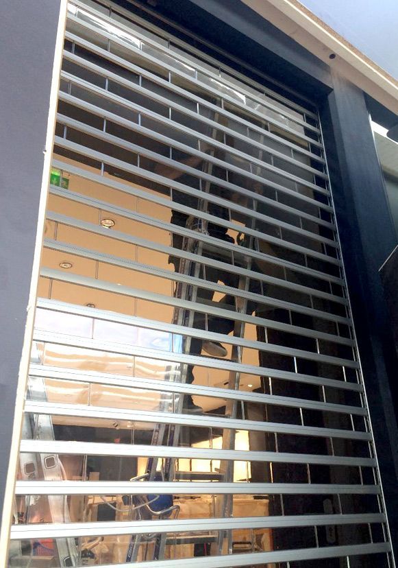 Volet de sécurité en métal argenté partiellement fermé sur la devanture d'un magasin.