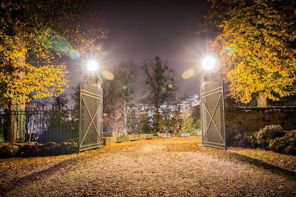 Des grilles en fer ouvertes, illuminées de lumières vives, mènent à un sentier dans un parc nocturne aux couleurs d'automne.