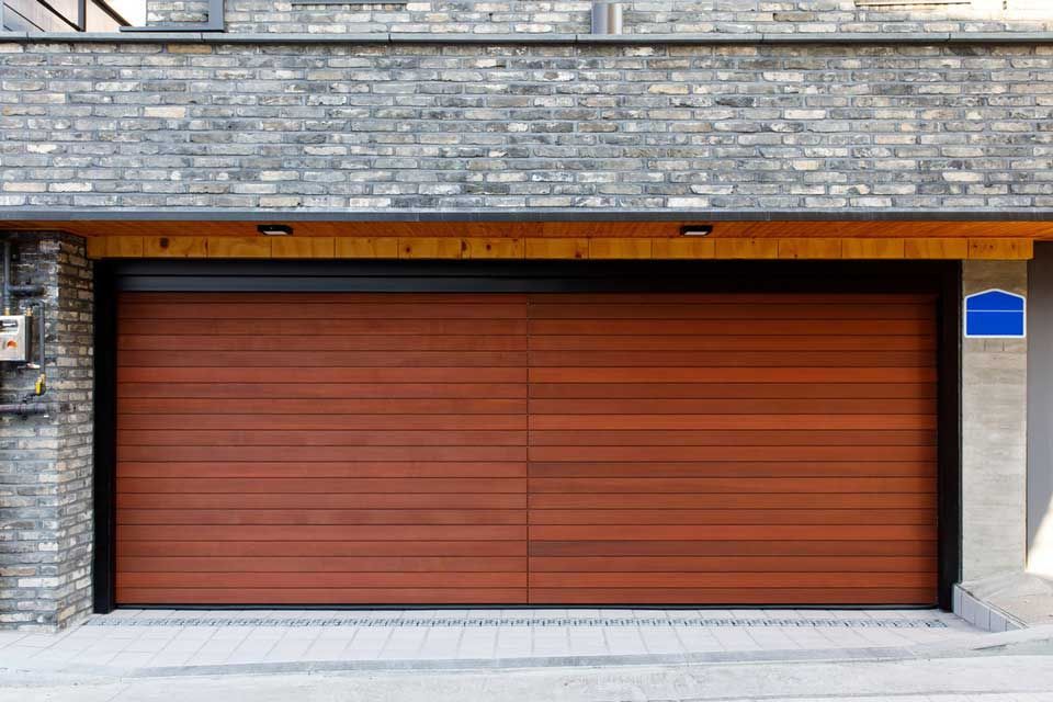 Porte de garage en bois brun sous un bâtiment en briques grises, allée pavée.