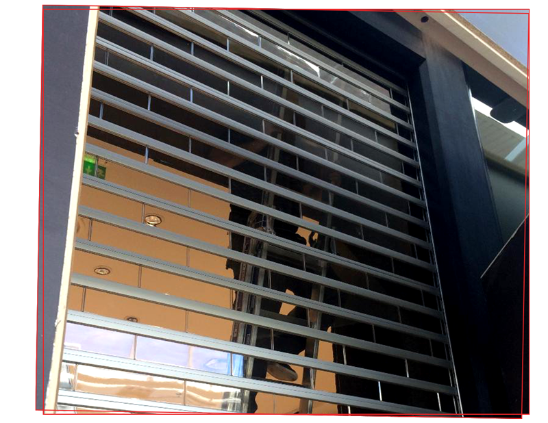 Grille de sécurité métallique, partiellement fermée, au-dessus d'une vitrine reflétant les bâtiments.