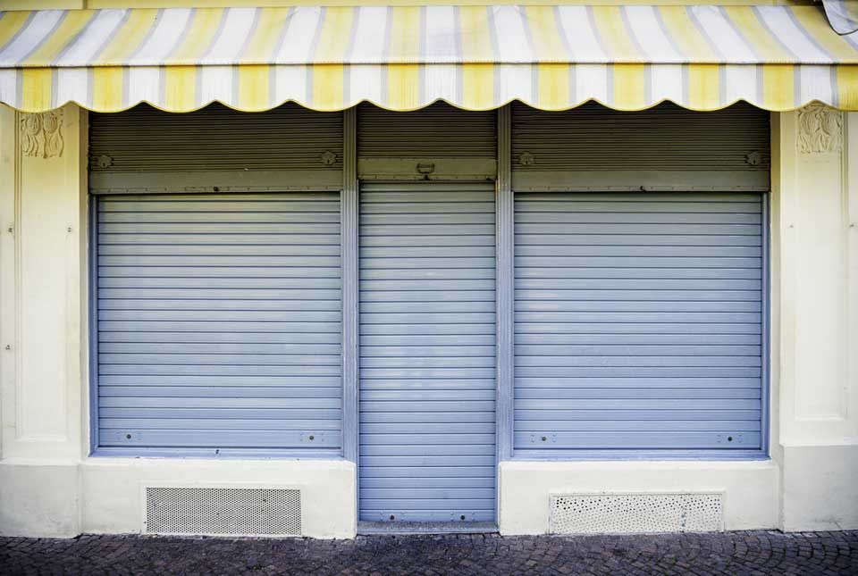 Façade fermée avec volets de sécurité en métal gris et auvent à rayures jaunes et blanches.