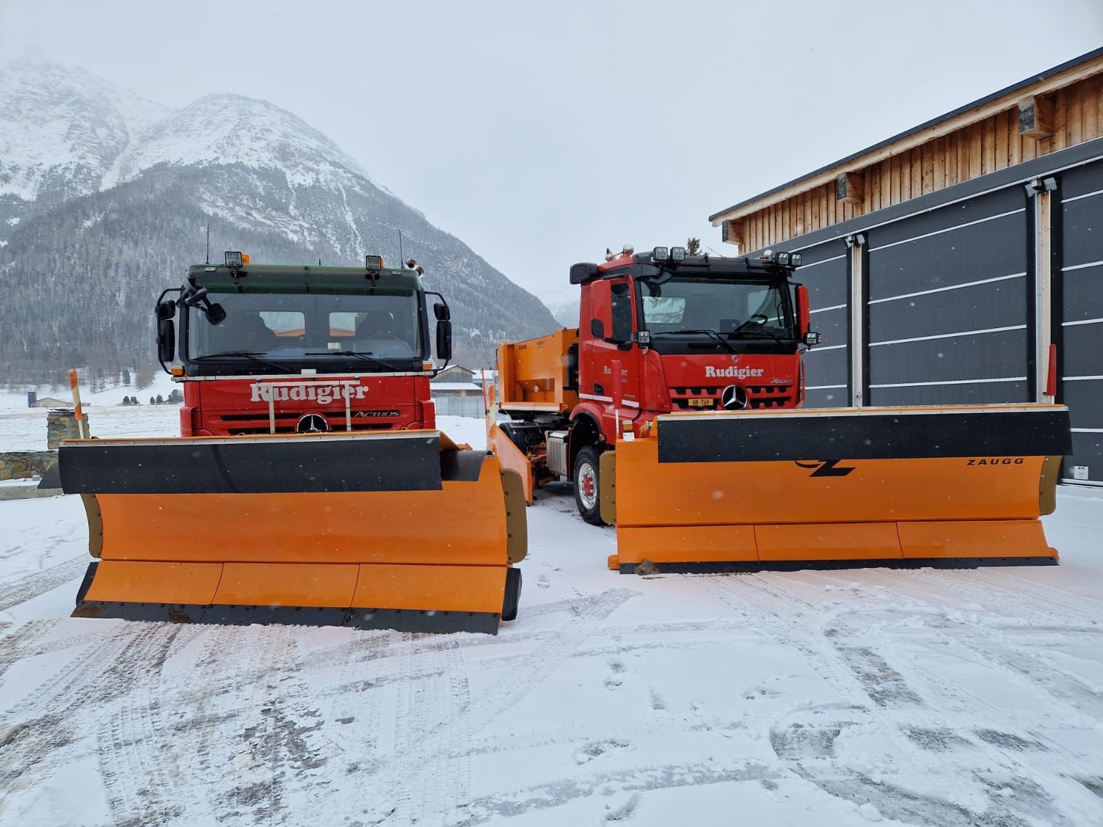 Ein gelber Schneepflug transportiert eine grosse Schneekugel auf einer verschneiten Strasse; im Hintergrund sind Berge zu sehen.